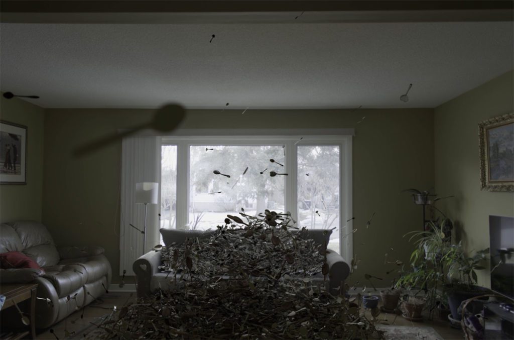 Image description: A photograph of a living room. The room is filled with hundreds of spoons that are seemingly falling from the ceiling, or exploding upwards from a pile on the floor. The room is dimly lit. The only light comes from the large picture window, which shows a snowy outdoor scene.